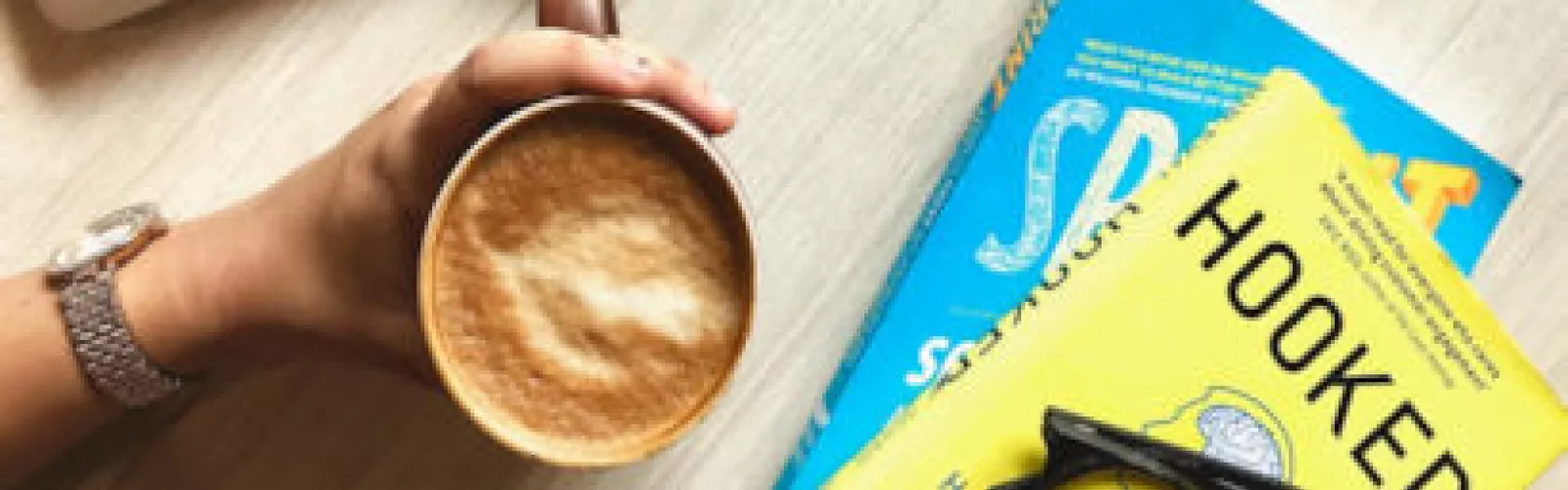 A hand holding a cup of frothed coffee next to two books lying flat on a table